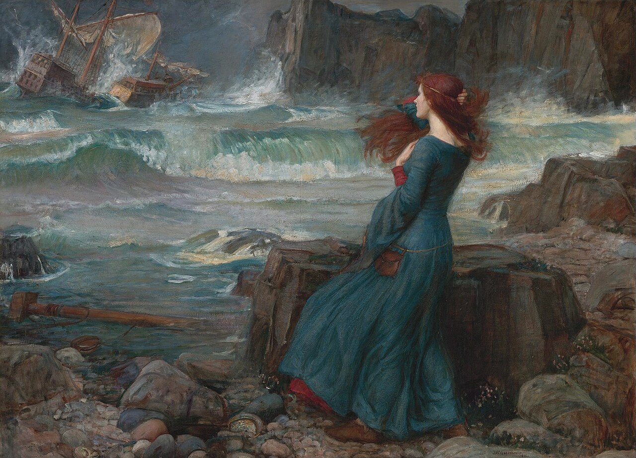 A rocky shore under a grey storm sky. A woman in a deep teal-blue dress stands with her back mostly to us on the pebbles, long red hair streaming sideways in the wind, one hand raised to hold it clear of her face as she watches the sea. Out to the left a three-masted ship is going over in heavy green-white breakers. Dark cliffs close the scene on the right; a broken spar and small pink flowers lie among the stones in the foreground.